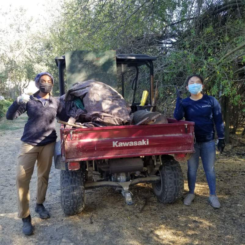 Two students with surgical masks on show a thumbs up near a tractor cart with bagged trash and debris cleaned up from the Woods area in a recent workparty. 