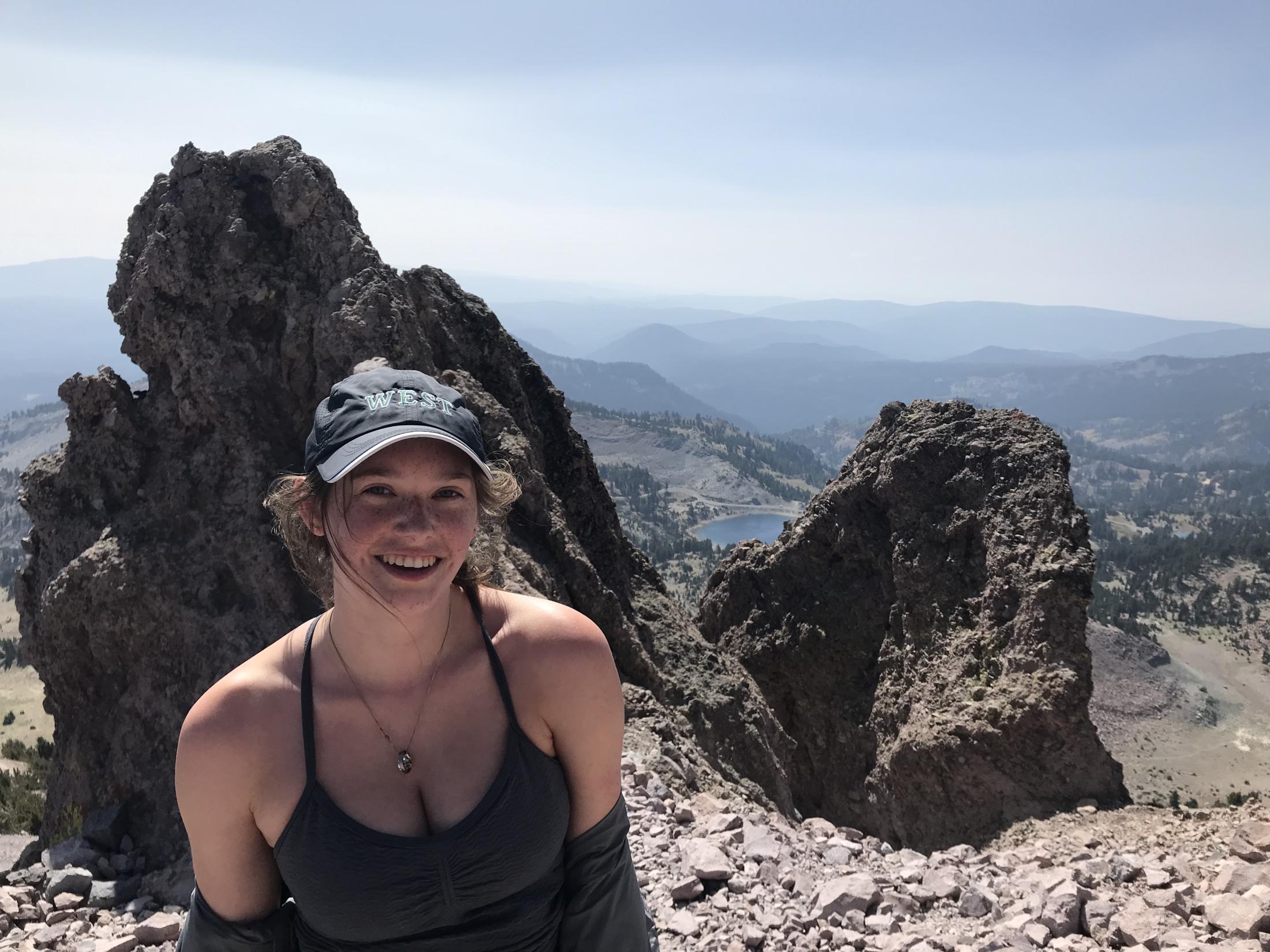 green fellow avery williamson poses on a windy mountain in a hat and tank top with the view of the blue horizon behind her