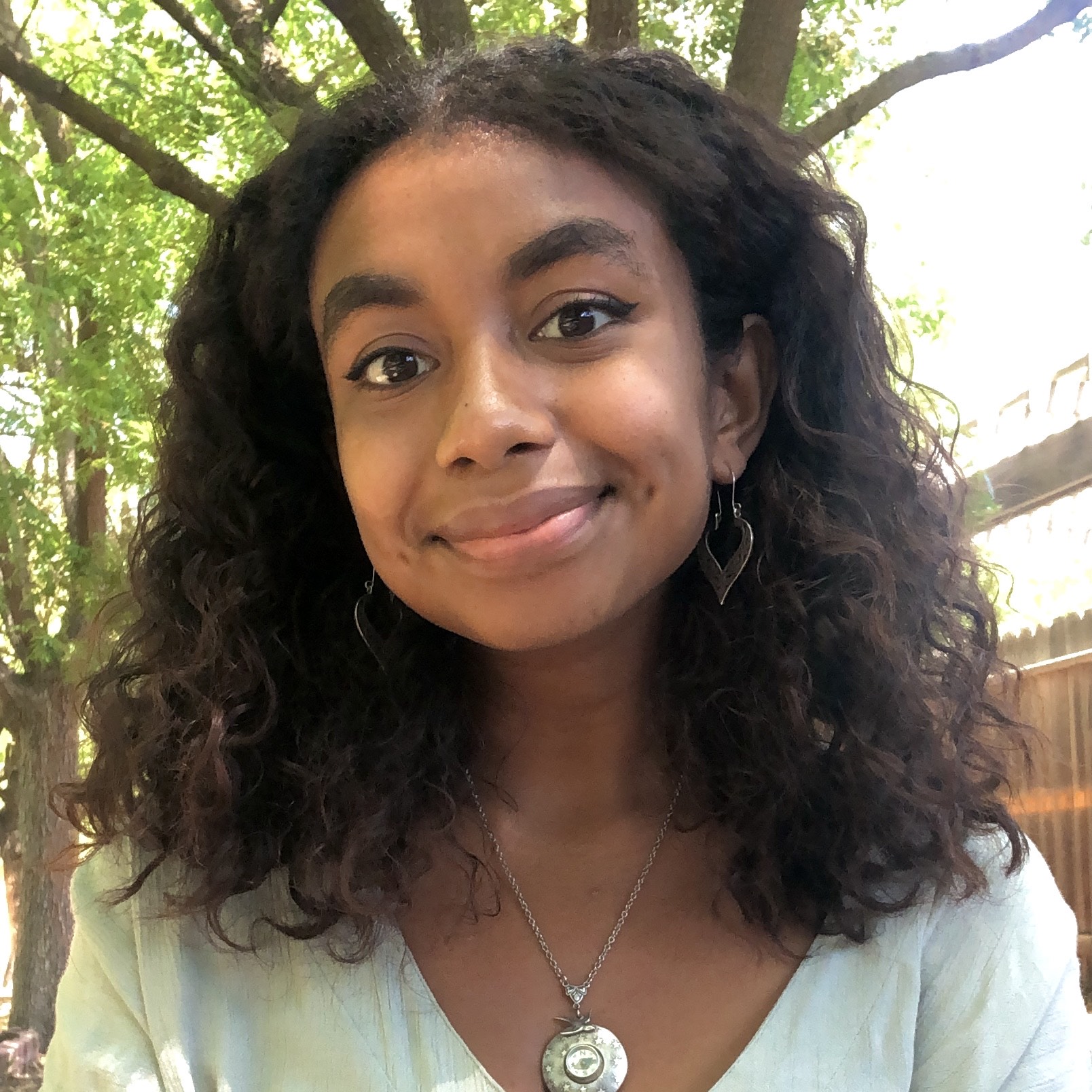 fellow suraya akhenaton smiling, wearing a light blue top and necklace under a tree outside